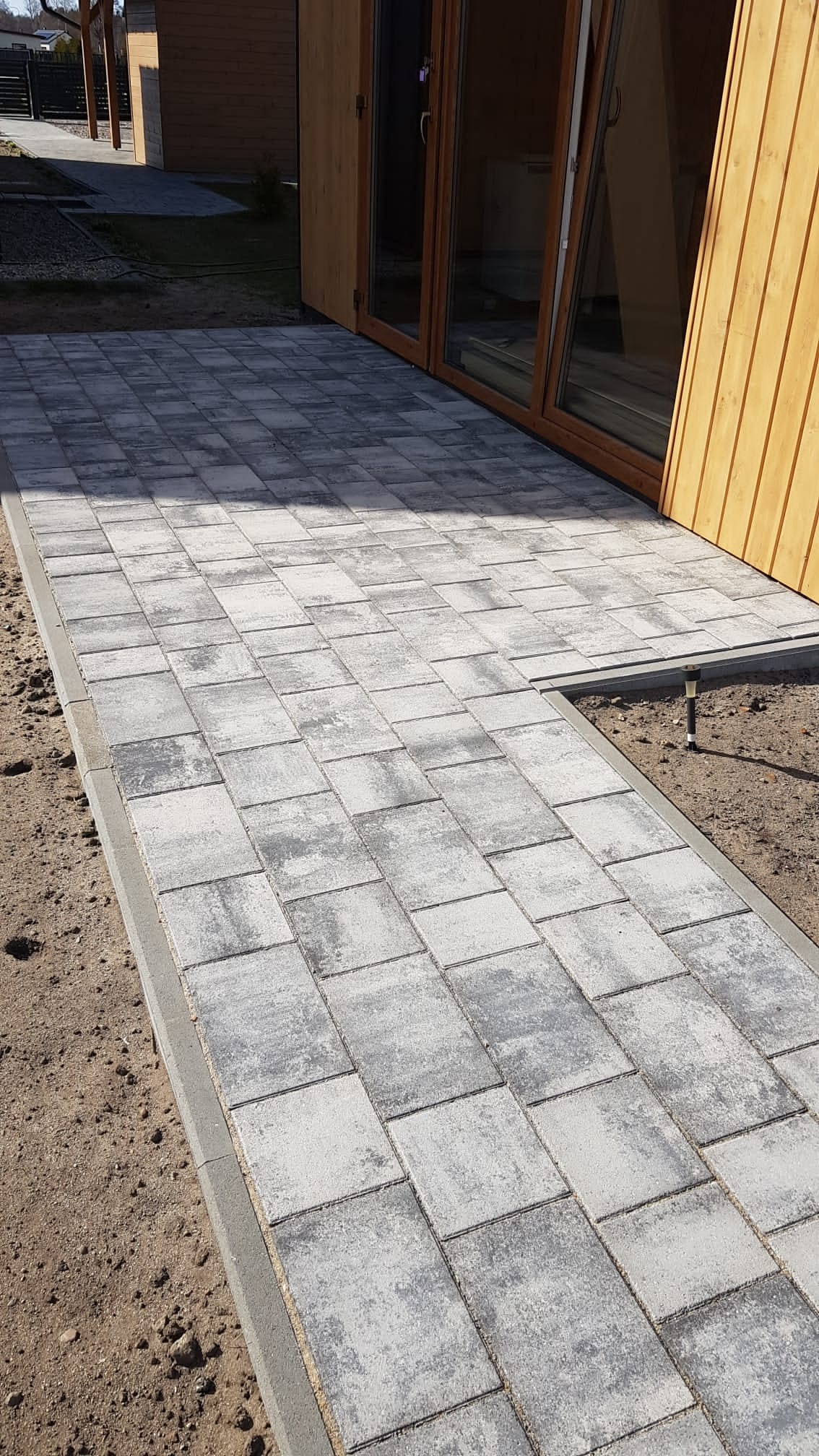 Ułożona szara kostka brukowa przed drewnianym domkiem, z widocznym odpływem liniowym i niezagospodarowanym terenem wokół.