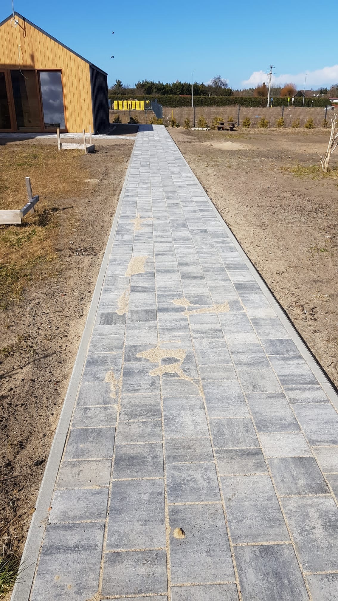 Długa, prosta ścieżka z szarej kostki brukowej prowadząca od drewnianego budynku w stronę ogrodzenia z siatki, widoczne pozostałości piasku na powierzchni kostki.
