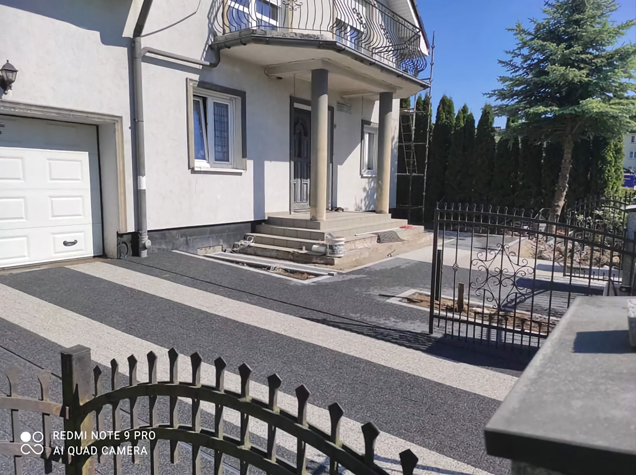 Dwukolorowa, pasiaste ułożona nawierzchnia przed domem z kolumnami, widoczne przygotowane podłoże pod dalszą część nawierzchni, czarny ozdobny płot w tle.