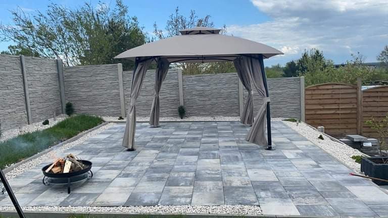 Szara kostka brukowa ułożona na tarasie z altaną i paleniskiem. W tle betonowe panele ogrodzeniowe i drewniany płot.