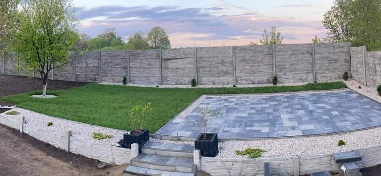 Ukończony taras z szarej kostki, otoczony białym żwirem i niskim murkiem, z trawnikiem i drzewem w tle, całość ogrodzona betonowym płotem o fakturze drewna.