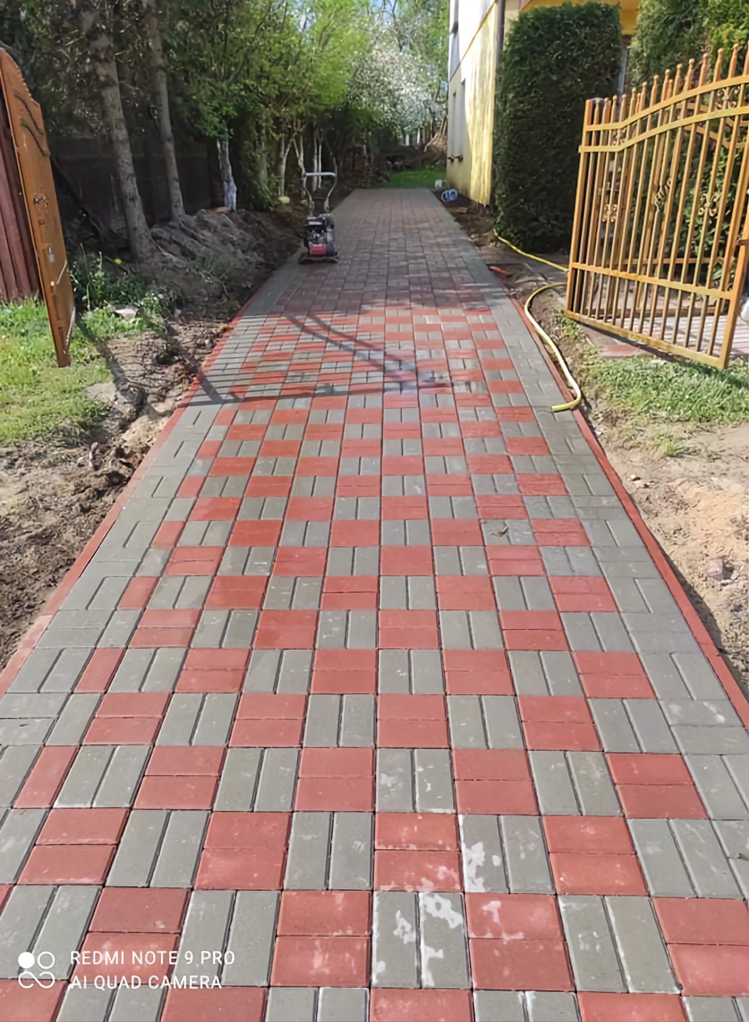 Nowo ułożony chodnik z czerwono-szarej kostki, wzór szachownicy, ubijarka w tle, widoczne świeże obsypanie ziemią po bokach.