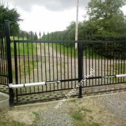 Czarna, metalowa brama wjazdowa z automatyką, widoczne siłowniki, na podjeździe kostka ażurowa, w tle zieleń i droga gruntowa.