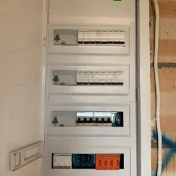 Otwarta skrzynka rozdzielcza z bezpiecznikami i wyłącznikami różnicowoprądowymi, widoczne przewody wewnątrz, zamontowana na ceglanej ścianie z niebieskimi plamami farby.