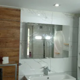 Nowoczesna łazienka z białymi marmurkowymi płytkami, drewnopodobnymi panelami na ścianie, białą szafką z drewnianymi uchwytami i dużym lustrem, w którym odbija się oświetlenie i fragment okna.