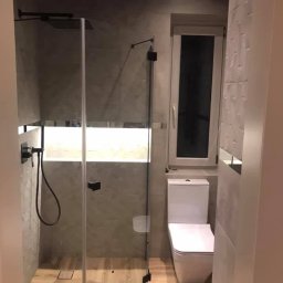 Usługi remontowe wykończenia wnętrz - Nowoczesna łazienka z kabiną prysznicową typu walk-in, drewnopodobną podłogą i szarą mozaiką na ścianach. Widoczna toaleta i okno. Minimalistyczny design.