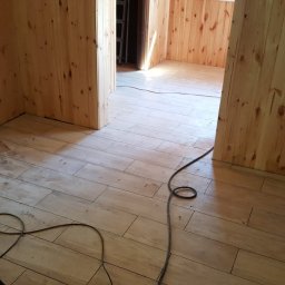 Usługi remontowe wykończenia wnętrz - Wykończone wnętrze z drewnianą boazerią i ułożoną podłogą z płytek imitujących drewno. Widoczne ślady po pracach wykończeniowych i kabel na podłodze.