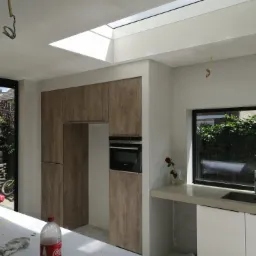 Wnętrze kuchni w trakcie prac wykończeniowych: świeżo położone gładzie, surowe wnęki okienne, okno dachowe, zabudowa meblowa z fornirem w kolorze drewna, widoczne przewody elektryczne, butelka...