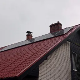 Panele fotowoltaiczne na dachu domu z czerwoną dachówką i dwoma kominami, widok z dołu w pochmurny dzień.