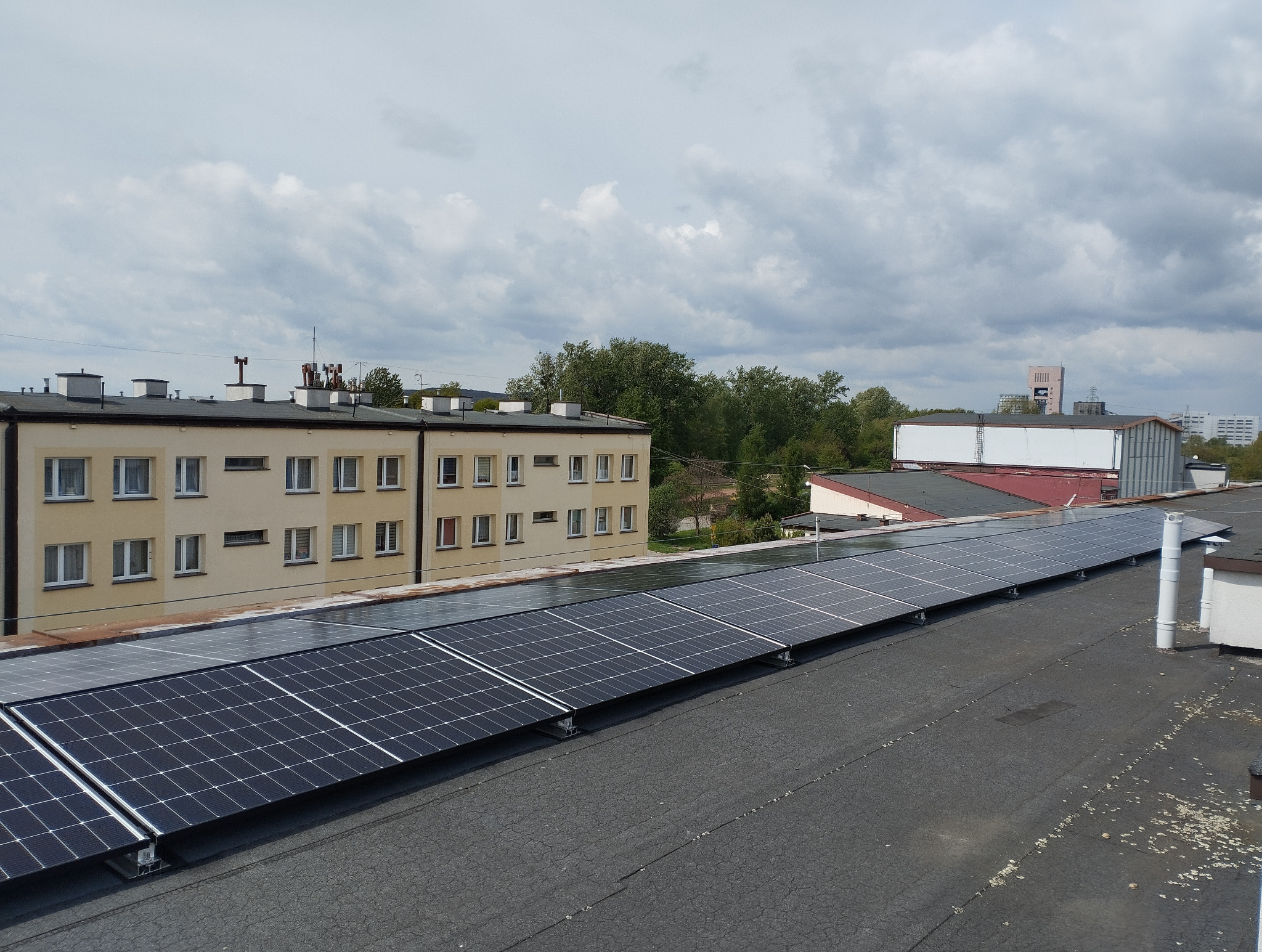 Rząd paneli fotowoltaicznych na płaskim dachu budynku, widok z góry, pochmurne niebo w tle. Widoczny fragment dachu, budynki mieszkalne i przemysłowe.