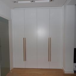 Sławmar Mebel Styl s.c. - Biała szafa wnękowa z czterema drzwiami i długimi, drewnianymi uchwytami, na podłodze z paneli, obok drzwi i kominka.