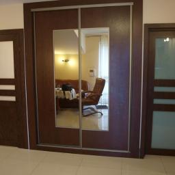 Sławmar Mebel Styl s.c. - Wnętrze z brązową szafą przesuwną z lustrem pośrodku, flankowaną przez dwie pasujące brązowe drzwi z szybami, na tle jasnych ścian i podłogi.