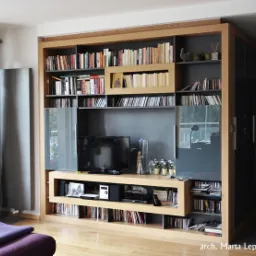 Zabudowa ściany w salonie z półkami na książki, szafkami z lakierowanymi frontami i miejscem na telewizor, wykonana z drewna w jasnym odcieniu dębu.