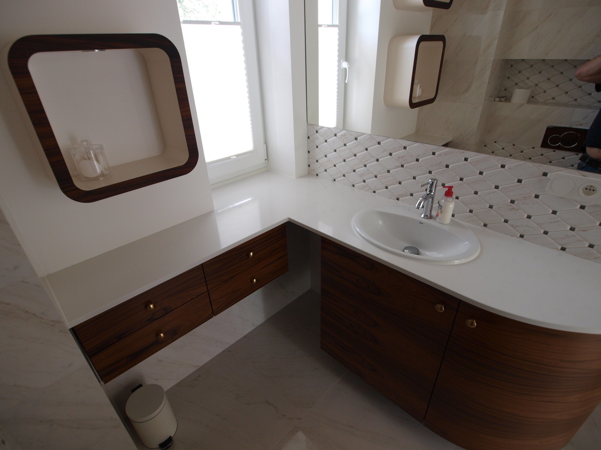 Nowoczesna łazienka z blatem zintegrowanym z umywalką, szafkami z ciemnego drewna i otwartą półką w ramie z tego samego drewna, uzupełniona marmurkową mozaiką na ścianie.