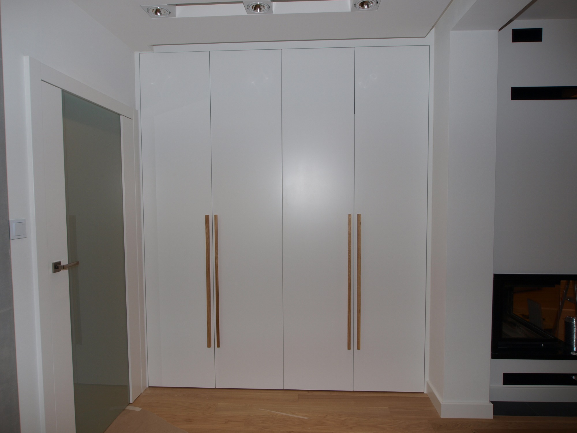 Biała szafa wnękowa z czterema drzwiami i długimi, drewnianymi uchwytami, na podłodze z paneli, obok drzwi i kominka.