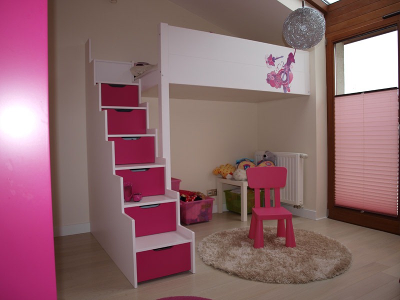 Białe łóżko piętrowe dla dziecka z różowymi akcentami, schody z szufladami, różowy stołek na okrągłym dywanie, dekoracja z jednorożcem na boku łóżka.