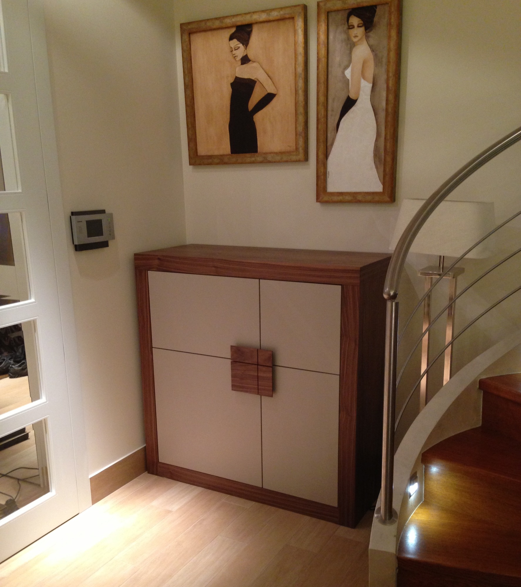 Nowoczesna szafka z drewnianymi elementami i kwadratowym uchwytem, umieszczona pod obrazami w eleganckim wnętrzu z drewnianymi schodami.