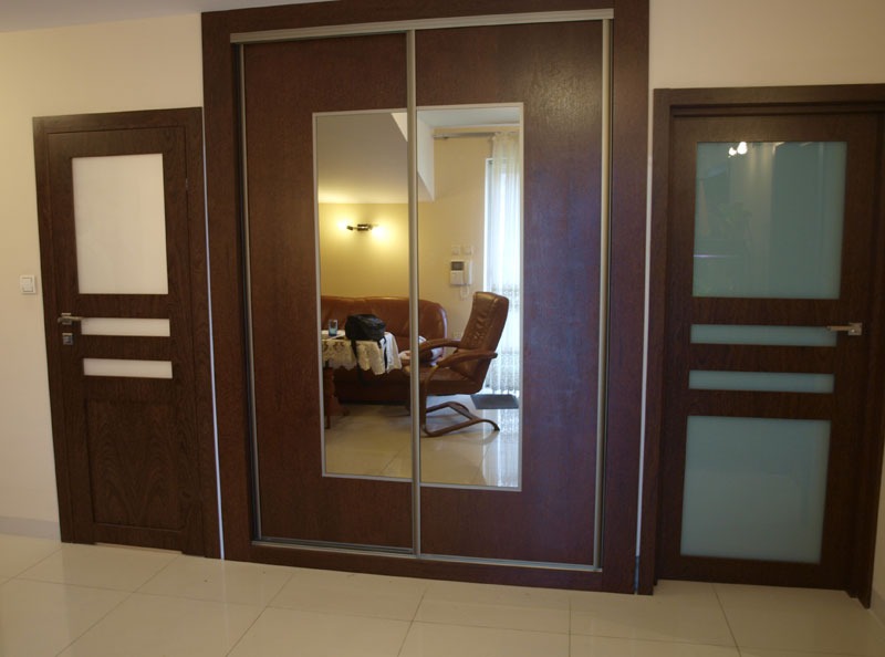 Wnętrze z brązową szafą przesuwną z lustrem pośrodku, flankowaną przez dwie pasujące brązowe drzwi z szybami, na tle jasnych ścian i podłogi.