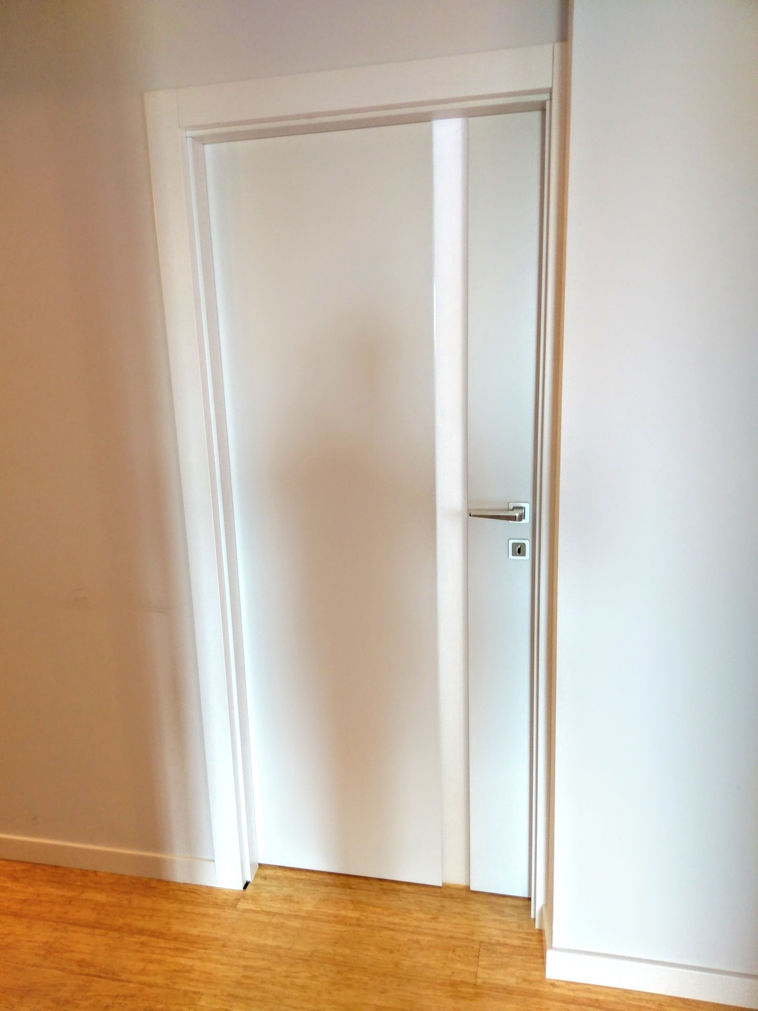 Białe drzwi wewnętrzne z pionowym przeszkleniem, srebrna klamka, drewniana podłoga, minimalistyczny design.