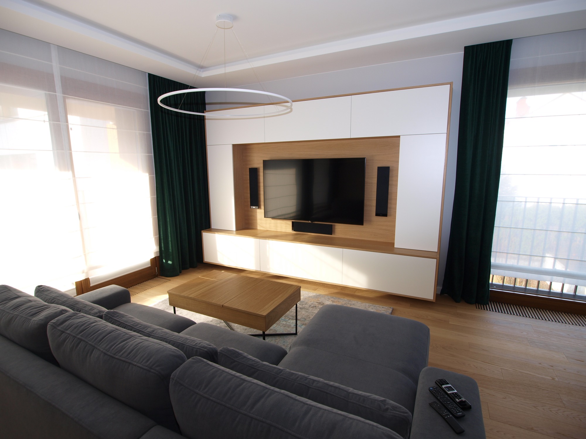 Nowoczesny salon z szarą sofą, drewnianym stolikiem kawowym i ścianą telewizyjną z białymi szafkami i drewnianym panelem, oświetlony okrągłą lampą wiszącą.