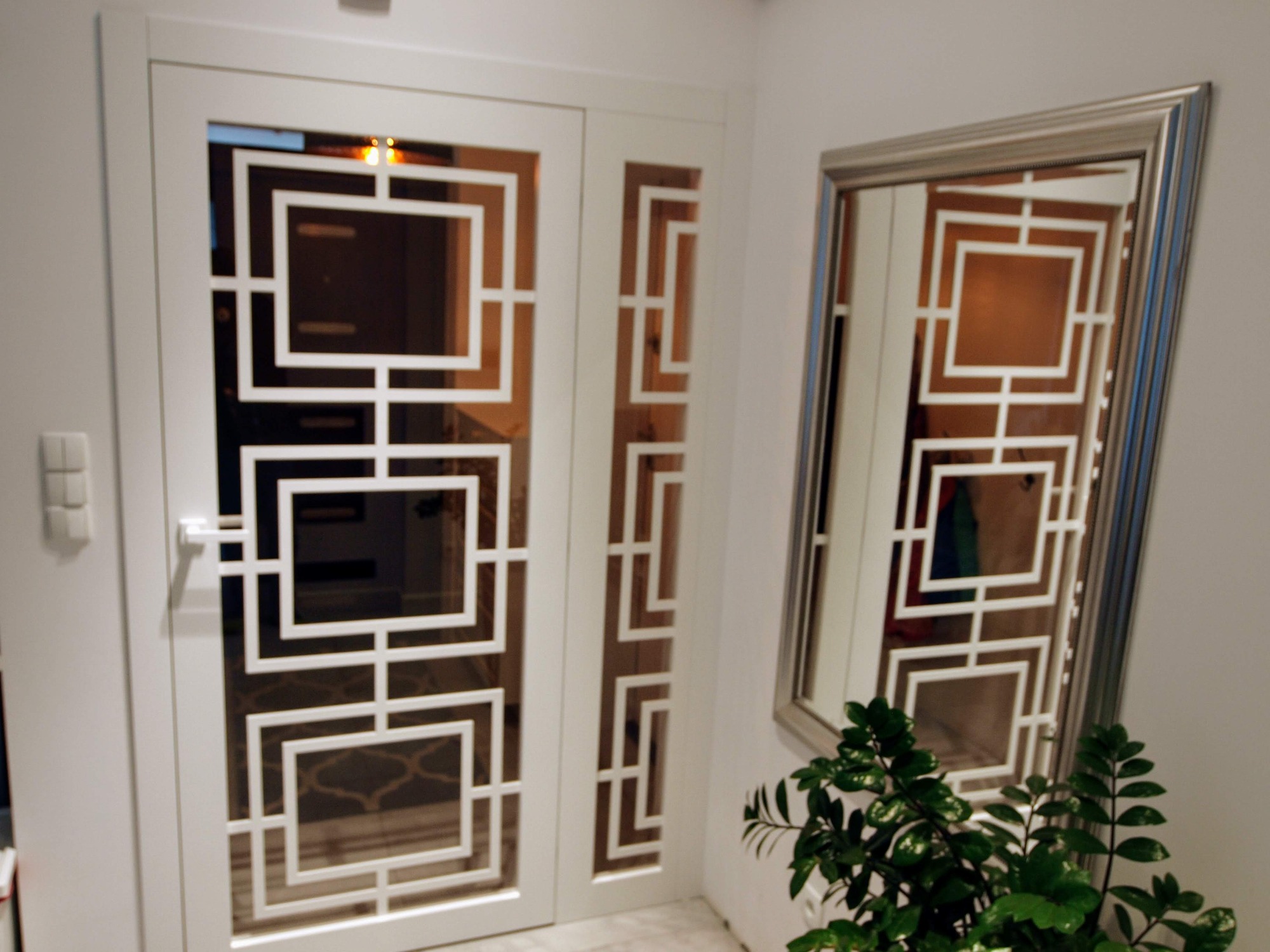 Białe drzwi przesuwne z geometrycznym wzorem kwadratów, obok lustro z podobnym motywem, uzupełnione zieloną rośliną w doniczce.