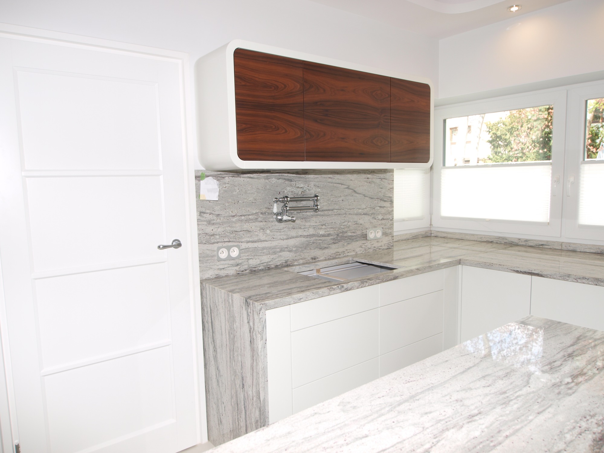 Nowoczesna kuchnia z białymi szafkami dolnymi i blatem z szarego kamienia z wyraźnym użyleniem, górna szafka fornirowana drewnem o ciepłym odcieniu i zaokrąglonych krawędziach, bateria ścienna...