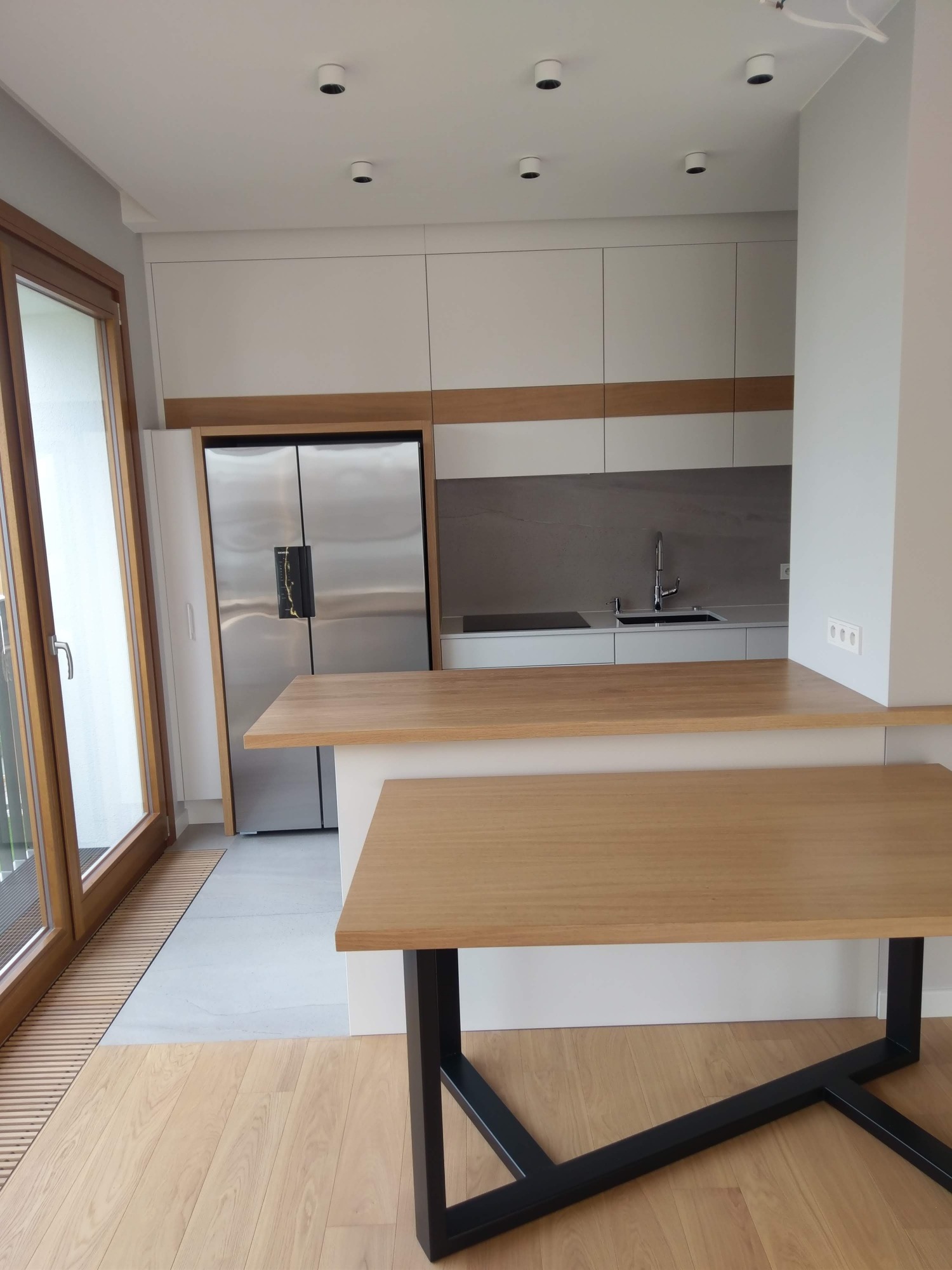 Nowoczesna kuchnia z białymi szafkami, drewnianym blatem i wyspą z czarnymi nogami, widok na okno balkonowe.