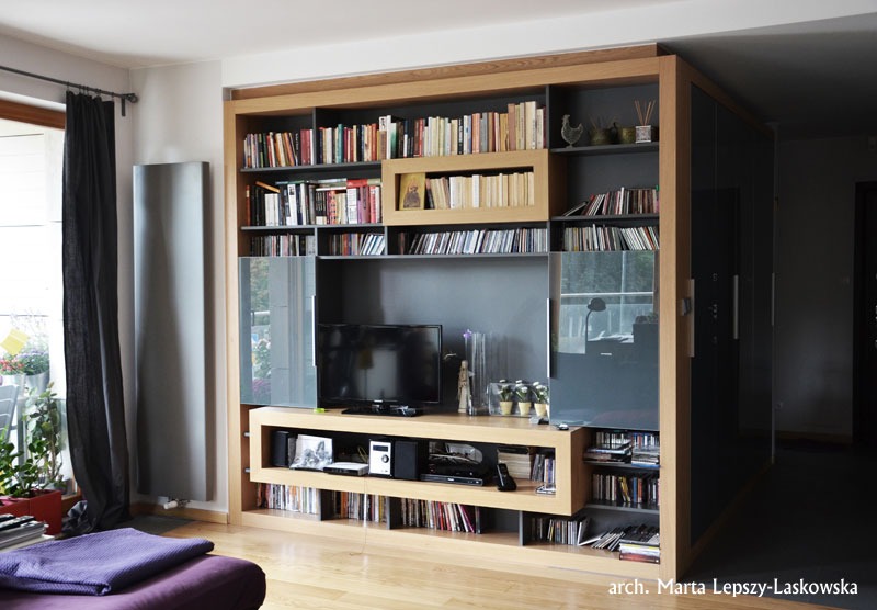 Zabudowa ściany w salonie z półkami na książki, szafkami z lakierowanymi frontami i miejscem na telewizor, wykonana z drewna w jasnym odcieniu dębu.