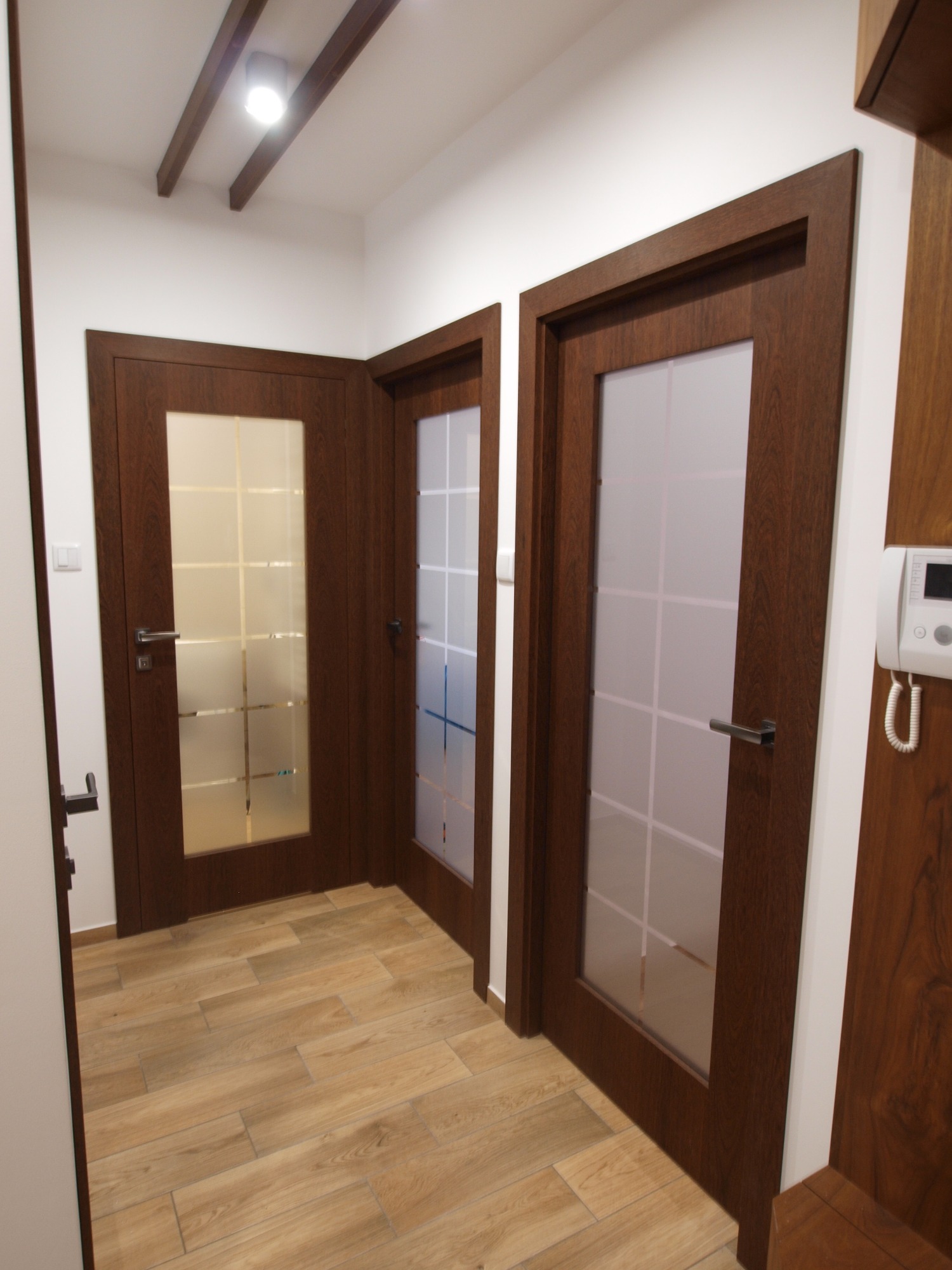 Trzy drewniane drzwi wewnętrzne z matowym szkłem w korytarzu, widoczny domofon i drewniane belki na suficie.