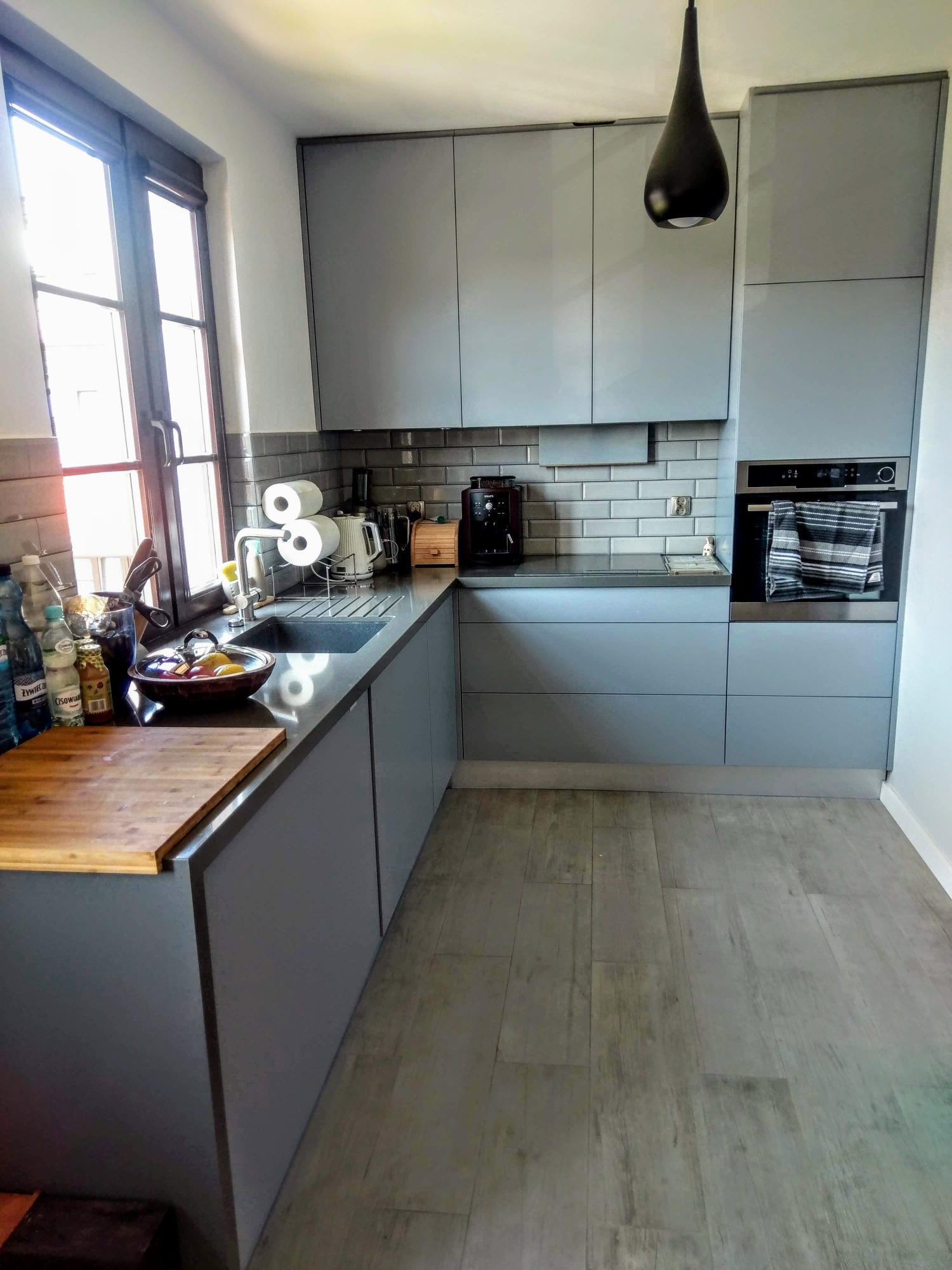 Szara kuchnia z drewnianym blatem, ciemnym zlewem i szarą glazurą na ścianie, z widocznym oknem i czarną lampą wiszącą.