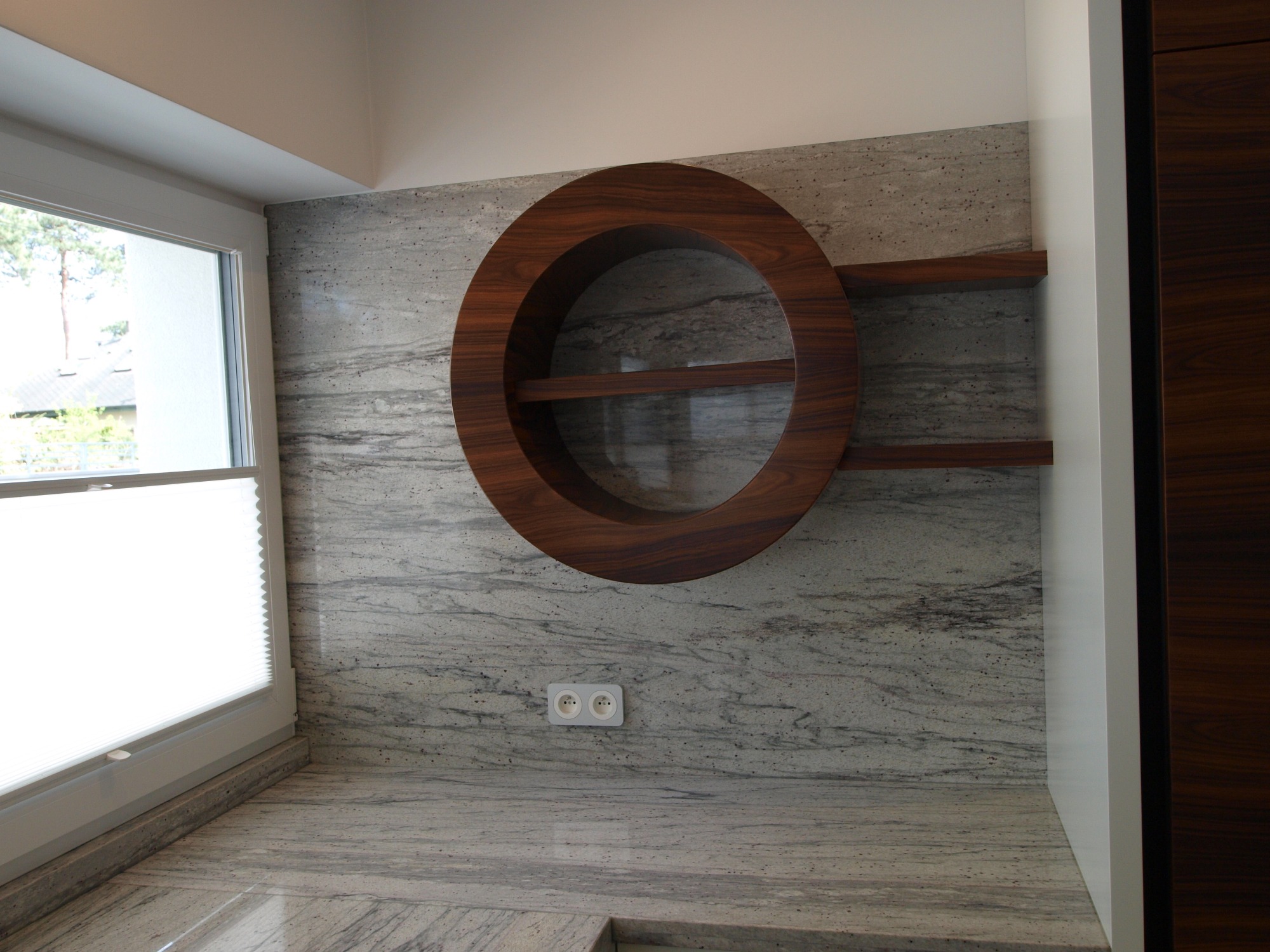 Okrągła, drewniana półka z dwoma poziomami wbudowana w kamienną ścianę, obok okno z roletą i fragment szafy.