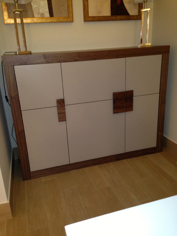 Nowoczesna komoda z beżowymi frontami i drewnianą ramą, zwieńczona dwoma lampkami, ustawiona na tle ściany z obrazami w złotych ramach.