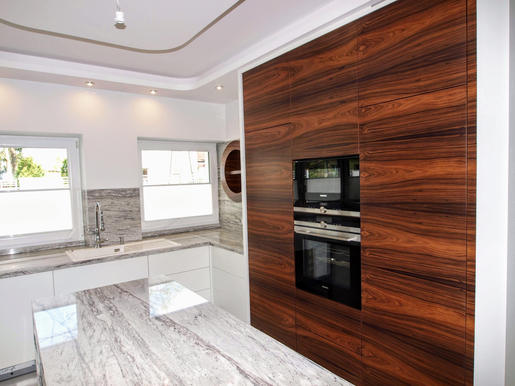 Nowoczesna kuchnia z wyspą i zabudową z forniru egzotycznego, zintegrowanymi piekarnikami Siemens i blatem z jasnego granitu z wyraźnym użyleniem.