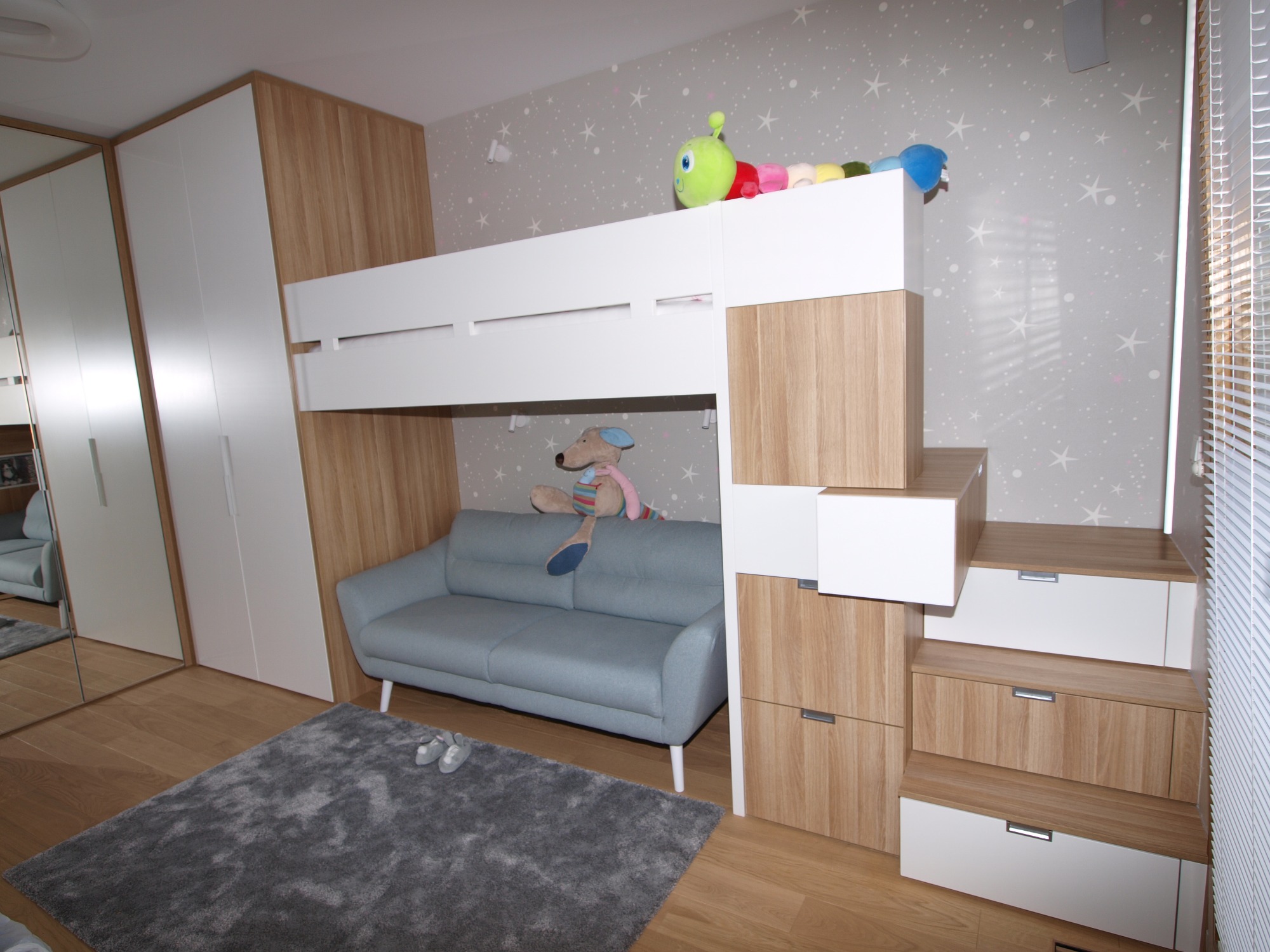 Pokój dziecięcy z zabudową: biało-drewniane łóżko piętrowe ze schodami z szufladami, szara sofa pod spodem, pluszaki na górze, lustrzana szafa obok, szary dywan na podłodze.