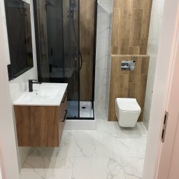 Grzegorz Rzeszot - Nowoczesna łazienka z kabiną prysznicową typu walk-in z czarnymi ramami, umywalką z szafką z drewnianym dekorem, podwieszaną toaletą i płytkami imitującymi drewno na ścianie.