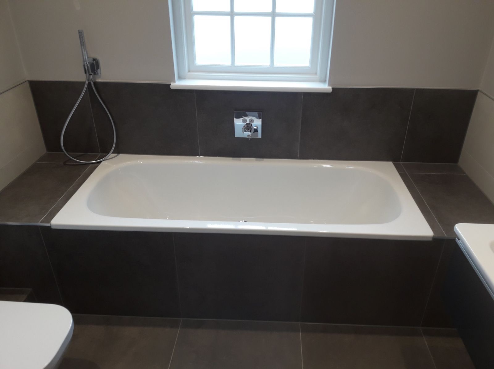 Biała wanna obudowana ciemnymi płytkami ceramicznymi pod oknem, bateria wannowa chromowana na ścianie, słuchawka prysznicowa na wężu, fragment toalety widoczny z lewej strony.