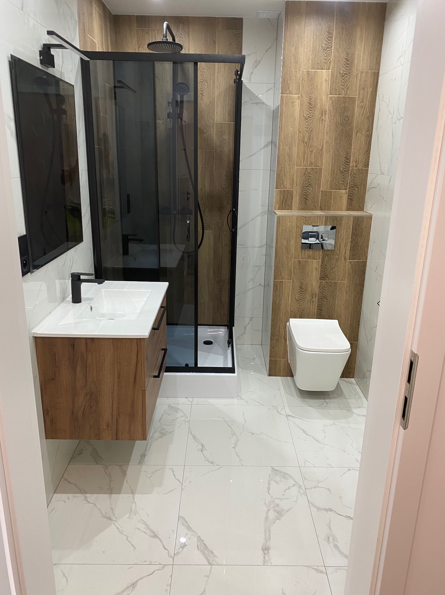 Nowoczesna łazienka z kabiną prysznicową typu walk-in z czarnymi ramami, umywalką z szafką z drewnianym dekorem, podwieszaną toaletą i płytkami imitującymi drewno na ścianie.