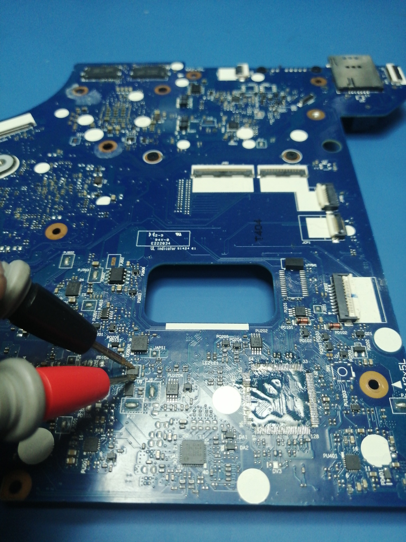 Niebieska płyta główna laptopa poddawana diagnostyce za pomocą multimetru z czarną i czerwoną sondą, widoczne liczne elementy SMD i układy scalone.