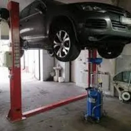 Szary samoch&oacute;d osobowy podniesiony na dwukolumnowym podnośniku hydraulicznym w warsztacie, widoczne urządzenie do spuszczania oleju, umywalka i fragment innego pojazdu w tle.