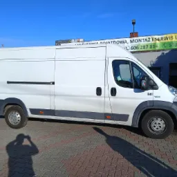 Biały samochód dostawczy zaparkowany przed budynkiem z banerem reklamującym montaż i serwis instalacji LPG i CNG.