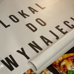 Plakat z napisem 'Lokal do wynajęcia' częściowo zwinięty w rulon, w tle widoczny fragment zdjęcia pizzy.
