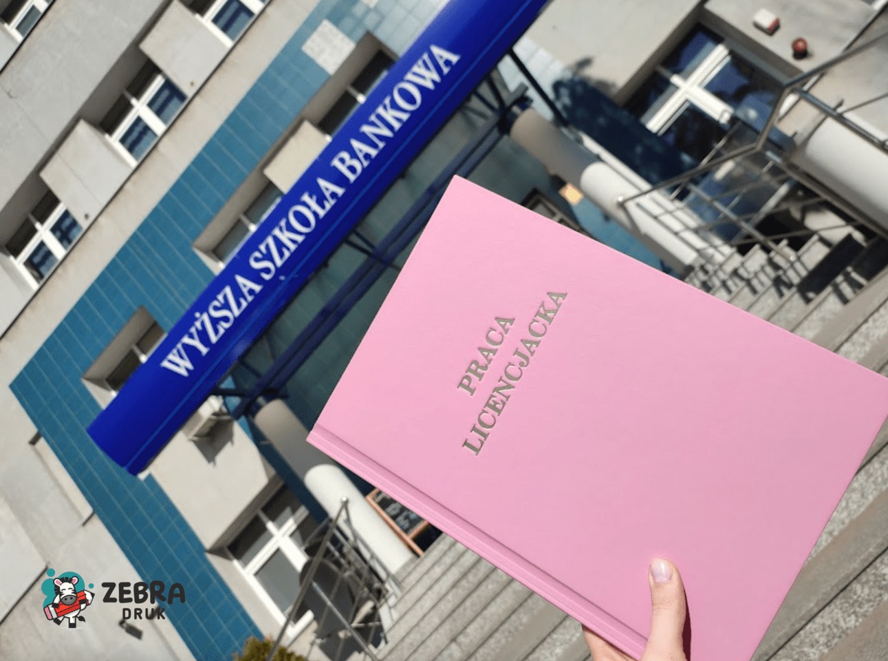 Różowa oprawiona praca licencjacka trzymana na tle budynku Wyższej Szkoły Bankowej, widoczny znak graficzny drukarni w lewym dolnym rogu.