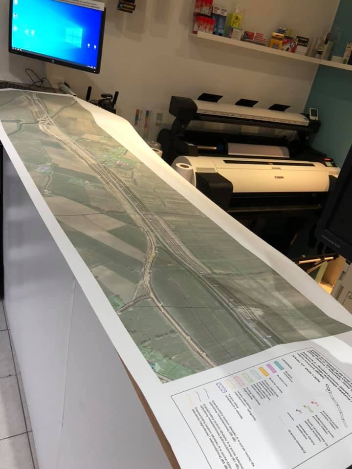 Duży wydruk mapy satelitarnej z naniesionymi oznaczeniami planowanej drogi, leżący na białym blacie, w tle widoczny ploter drukujący i monitor.