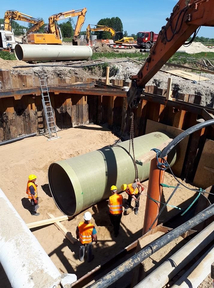 Montaż rury kanalizacyjnej w wykopie zabezpieczonym stalowymi ściankami; pracownicy w kamizelkach odblaskowych i kaskach nadzorują proces, w tle widoczne koparki i ciężarówka.