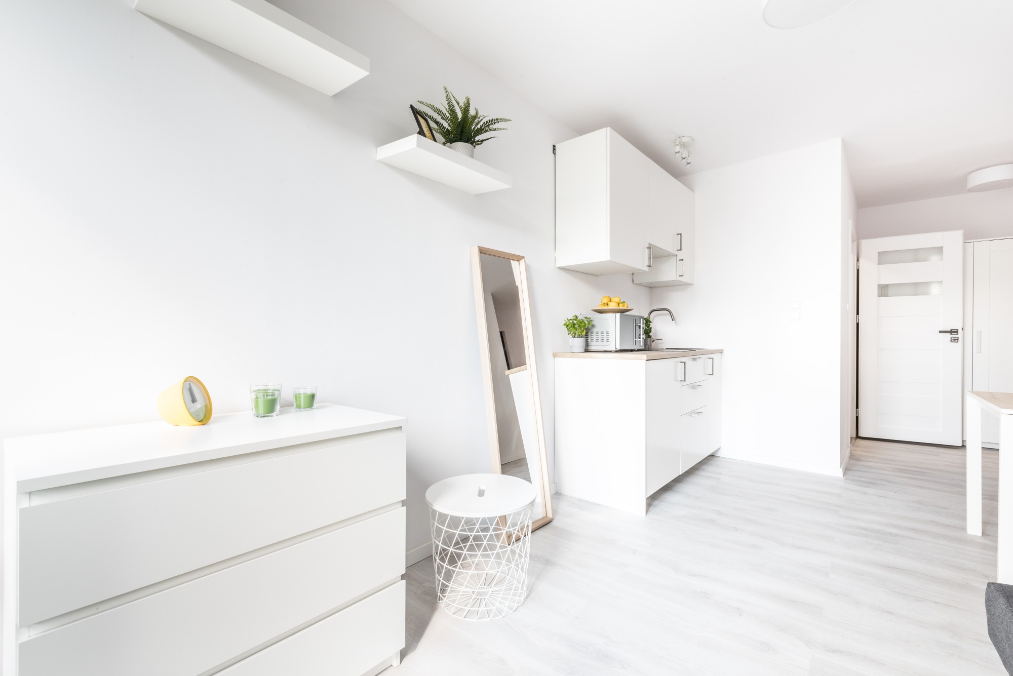 Jasne, minimalistyczne wnętrze z białymi meblami, drewnianą podłogą i akcentami dekoracyjnymi, takimi jak zegar, roślina doniczkowa i lustro stojące.