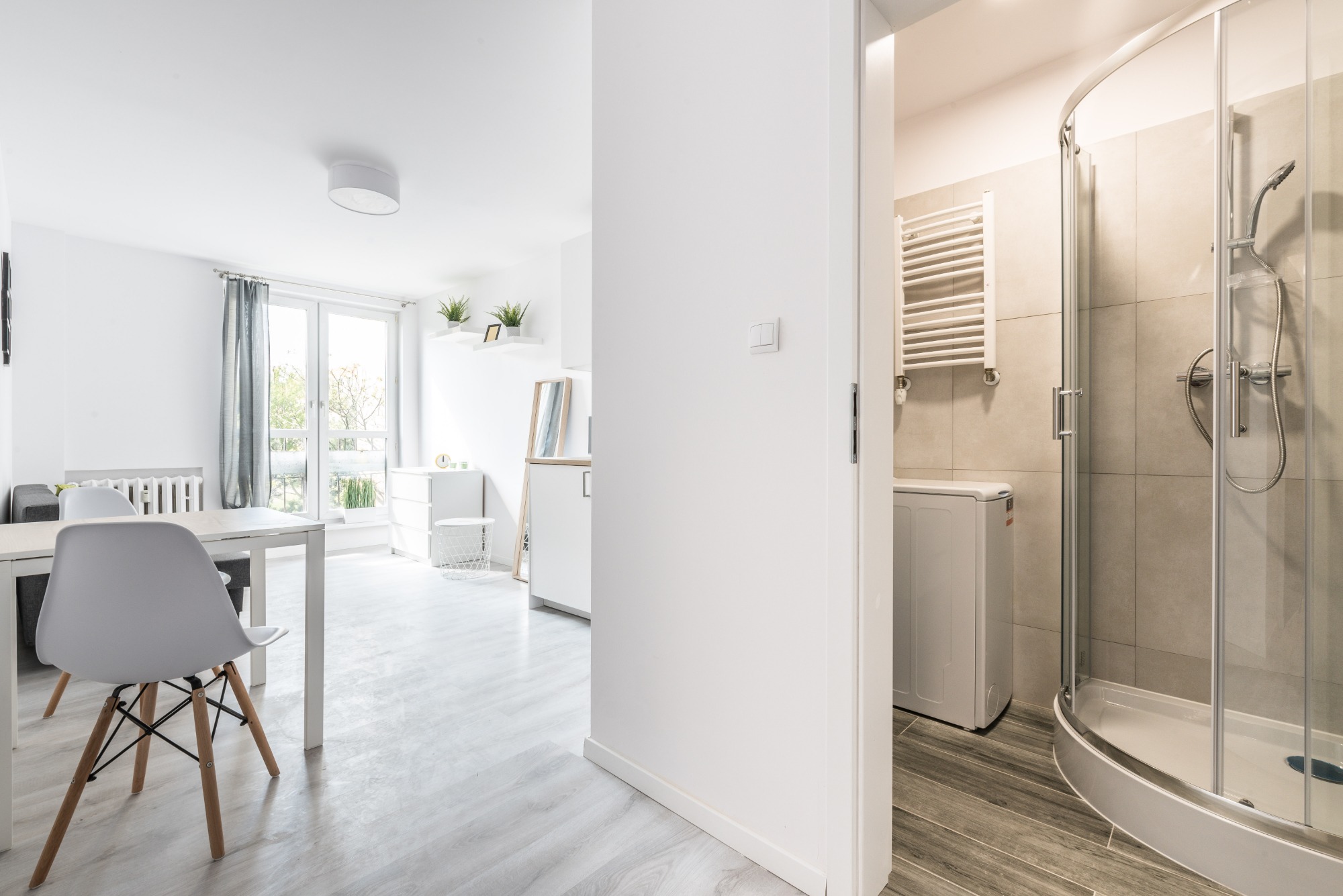 Widok z salonu na łazienkę z kabiną prysznicową i pralką, białe wnętrze z drewnianymi akcentami i szarymi płytkami.