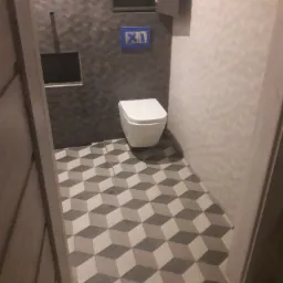 Wykończona łazienka z szaro-białymi płytkami podłogowymi tworzącymi iluzję 3D, szare płytki ścienne z drobnym wzorem, biała wisząca toaleta, zabudowana szafka nad toaletą, widok zza framugi drzwi.