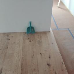 SzymańscybudPL - Układanie jasnej drewnianej podłogi, widoczna niebieska taśma malarska na zabezpieczonej podłodze w tle i zielona zmiotka przy ścianie.