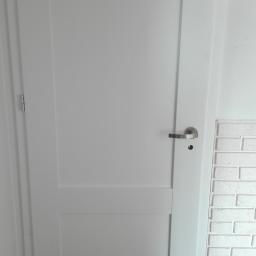 SzymańscybudPL - Białe, dwupanelowe drzwi wewnętrzne z srebrną klamką i widoczną ścianą wykończoną cegłą dekoracyjną.