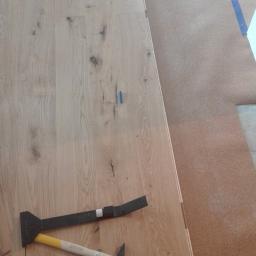 SzymańscybudPL - Montaż drewnianej podłogi: widoczne panele, podkład korkowy, niebieska taśma malarska, dobijak i młotek.